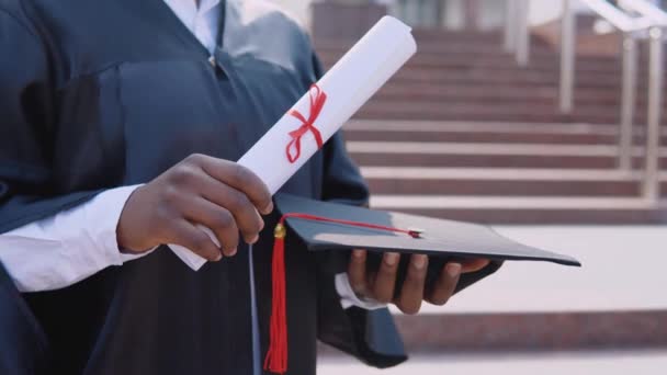 Kwadratowy kapelusz mistrzowski i dyplom ukończenia studiów w rękach Afroamerykanki absolwentki uniwersytetu na tle schodów z zewnątrz. — Wideo stockowe