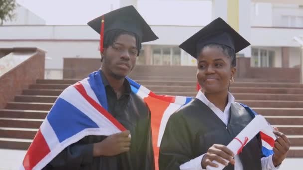 African American man and woman stand side by side facing the camera in black robes and square hats of graduate students with the British flag on their shoulders — Stock Video