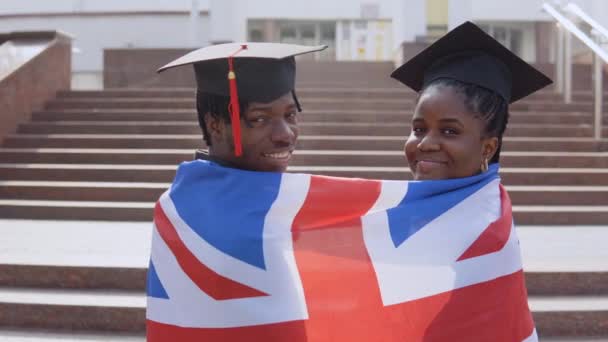 Hombre y mujer afroamericanos de pie lado a lado con sus espaldas a la cámara con túnicas negras y sombreros cuadrados de estudiantes graduados con la bandera británica sobre sus hombros. Mirando a la cámara — Vídeo de stock