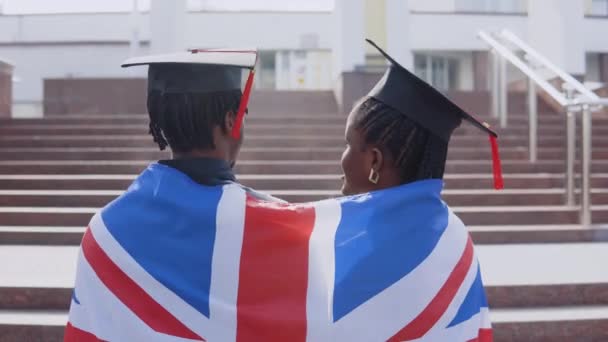 Afrikai amerikai férfi és nő egymás mellett állnak háttal a kamerának.A brit zászló a vállukon van. A háttérre épülő egyetemi épület — Stock videók