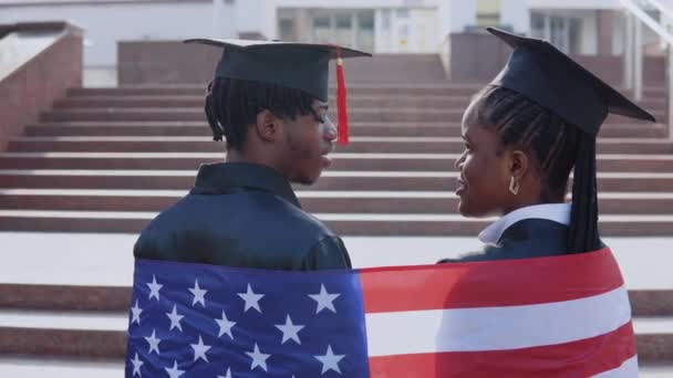 Афроамериканец и женщина стоят бок о бок спиной к камере. У них на плечах британский флаг. Здание университета на заднем плане — стоковое видео