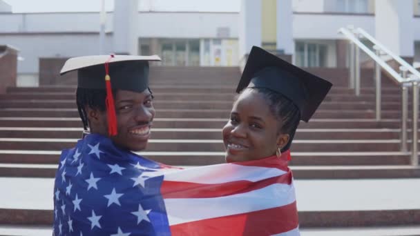 Hombre y mujer afroamericanos de pie lado a lado con la espalda a la cámara en sombreros cuadrados de estudiantes graduados con la bandera de Estados Unidos sobre sus hombros. Mirando a la cámara — Vídeo de stock