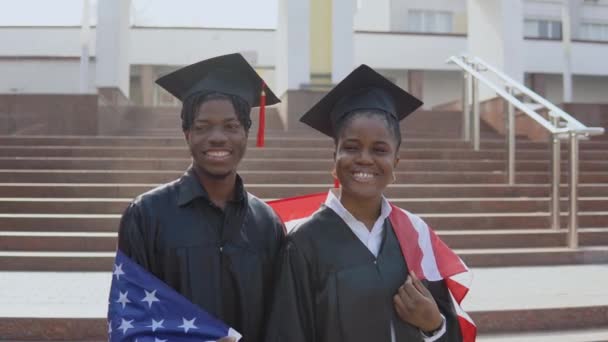 Afroamerykanin mężczyzna i kobieta stoją ramię w ramię twarzą do kamery w czarnych szatach i kwadratowych kapeluszach absolwentów z flagą USA na ramionach — Wideo stockowe