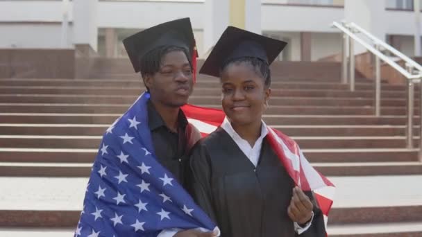 Αφροαμερικάνος άνδρας και γυναίκα στέκονται δίπλα-δίπλα αντικρίζοντας την κάμερα με μαύρες ρόμπες και τετράγωνα καπέλα μεταπτυχιακών φοιτητών με τη σημαία των ΗΠΑ στους ώμους τους — Αρχείο Βίντεο