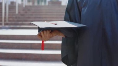Afro-Amerikan bir üniversite mezunu kadının elinde yatay olarak şapkalar var. Arka planda, merdivenlerin dışından..