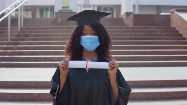 Młoda, afrykańska absolwentka, stojąca przed kamerą z dyplomem w rękach. Student ma ochronną maskę medyczną. Stoi na schodach na zewnątrz.. — Wideo stockowe