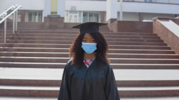 Una joven mujer afroamericana graduada se para frente a la cámara y se quita una máscara médica protectora de la cara. El estudiante está parado en las escaleras afuera.. — Vídeo de stock