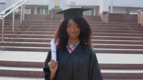 Молодая американка из Африки, выпускница, стоящая перед камерой с дипломом и книгами в руках. Студент стоит на лестнице снаружи. — стоковое видео