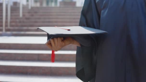 Sombrero de maestros cuadrados horizontalmente en las manos de una mujer afroamericana graduada en la universidad en el fondo de las escaleras desde el exterior. — Vídeo de stock