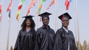 Üç Afro-Amerikan üniversite mezunu dünyanın dört bir yanından gelen bayrakların önünde duruyor. Öğrenciler üstat üniforması giymişler.