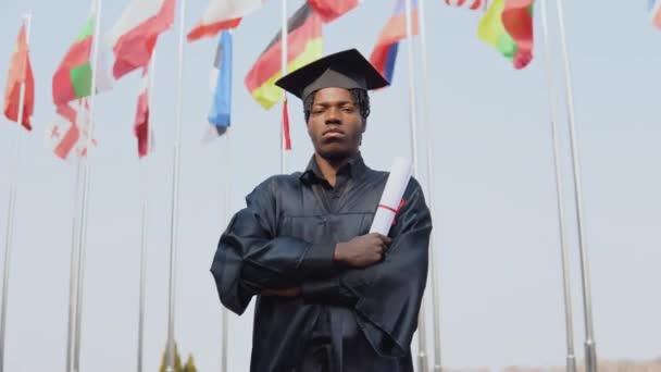 Jovem afro-americano graduado em pé em frente à câmera com um diploma em suas mãos. O estudante tem um chapéu de mestres quadrados e fica do lado de fora com as bandeiras internacionais no fundo. — Vídeo de Stock