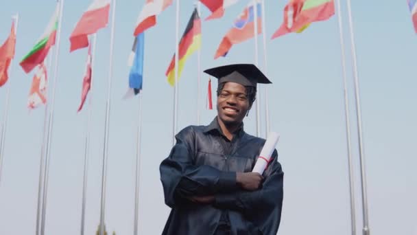 Feliz joven afroamericano graduado de pie delante de la cámara con un diploma en sus manos. El estudiante está afuera con las banderas internacionales en segundo plano. — Vídeo de stock