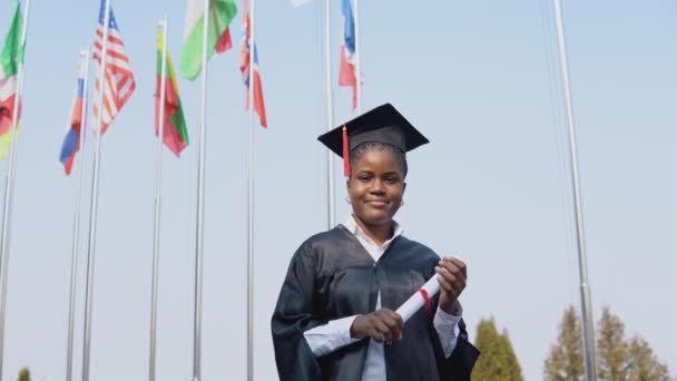 Молода афроамериканська випускниця стоїть перед камерою з дипломом у руках. Студент стоїть на вулиці з міжнародними прапорами на тлі. — стокове відео
