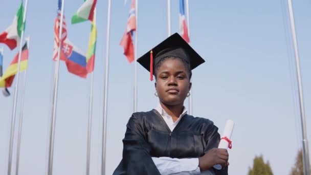 Jovem afro-americana graduada em pé em frente à câmera com um diploma em suas mãos. O estudante está lá fora com as bandeiras internacionais em segundo plano. — Vídeo de Stock