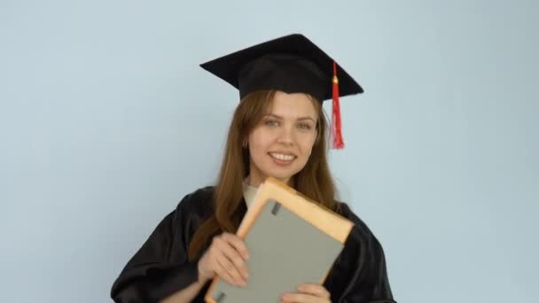 Молода студентка в чорному халаті і капелюх господарів трясе книжки, які вона тримає в руках. Білий фон — стокове відео