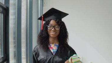 Genç Afrikalı Amerikalı kadın üniversite mezunu elinde kitaplarla siyah çerçeveli, geniş ve modern bir pencerenin yanında duruyor. Siyah cübbeli, kare şapkalı ve gözlüklü bir kız öğrenci.