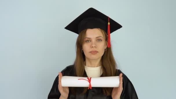 Młoda studentka w czarnej sukni i kapeluszu mistrzowskim trzyma w rękach w pozycji poziomej dyplom ukończenia studiów wyższych. Białe tło — Wideo stockowe