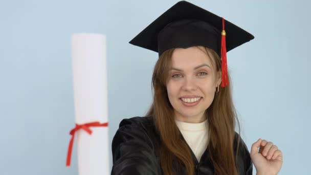 Jeune étudiante caucasienne en robe noire et chapeau de maître tient dans ses mains en position verticale un diplôme d'enseignement supérieur. Un diplômé de l'université danse avec un diplôme. Fond blanc — Video
