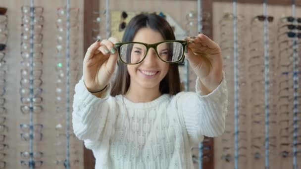 Eine junge Brünette mit langen Haaren steht in einem weißen Pullover vor einem Regal in einem Optikgeschäft und blickt durch eine Brille, die sie in ausgestreckten Armen hält. — Stockvideo