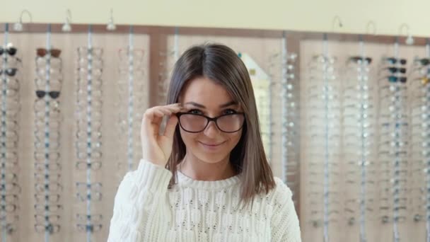 Ung brunett med blå ögon i en mysig vit tröja i en optikbutik drar upp glasögon — Stockvideo