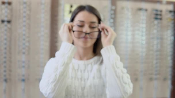 Una giovane bruna con gli occhi azzurri in un accogliente maglione bianco in un negozio di ottica tiene gli occhiali davanti alla telecamera e poi le mette gli occhiali sul viso — Video Stock