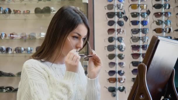 Wesoła młoda kobieta w przytulnym białym swetrze przymierza okulary przeciwsłoneczne w pobliżu zabytkowego lustra stołowego w salonie optycznym, a następnie patrzy w kamerę — Wideo stockowe