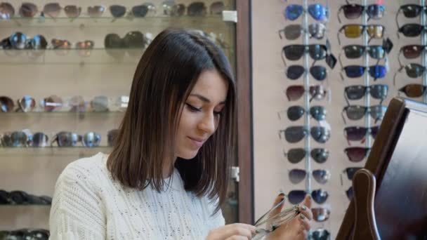 Fröhliche junge Kaukasierin im kuscheligen weißen Pullover probiert in der Nähe eines Vintage-Tischspiegels im Optiksalon eine durchsichtige Brille an und blickt dann in die Kamera — Stockvideo