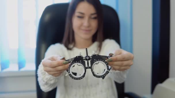 Een jonge vrouw in een knusse witte trui demonstreert aan de camera, en probeert dan skiascopische lenzen in de optische salon, kijkend naar de camera — Stockvideo