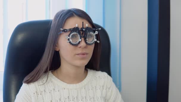 Egy fiatal nő egy kényelmes fehér pulóverben a szemész rendelőjében az optika klinikán válaszol egy kérdésre egy szemorvostól.. — Stock videók