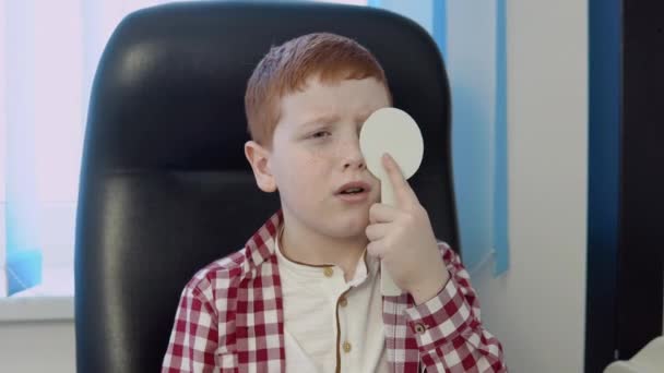 Een roodharige jongen in een rood-wit geruit shirt in het oogheelkundig kantoor in de optische kliniek beantwoordt een vraag van een arts tijdens een visie test. — Stockvideo