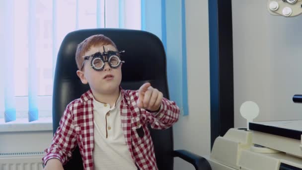 Un garçon roux avec des bretelles dans une chemise à carreaux rouge et blanc dans le bureau des ophtalmologistes à la clinique d'optique répond à une question des médecins lors d'un test de vision. — Video