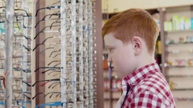 Ekose kırmızı ve beyaz gömlekli kızıl saçlı beyaz bir çocuk mercek çerçeveli bir askılığın yanında güneş gözlüğü deniyor. Yan görünüm