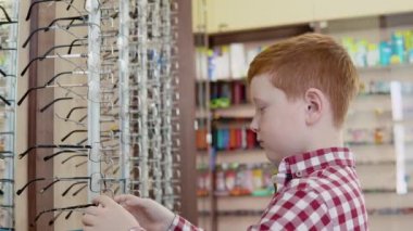 Ekose kırmızı ve beyaz tişörtlü kızıl saçlı bir çocuk askıdan bir çerçeve alır ve mercek çerçevesiyle askılığın yanında dener. Yan görünüm