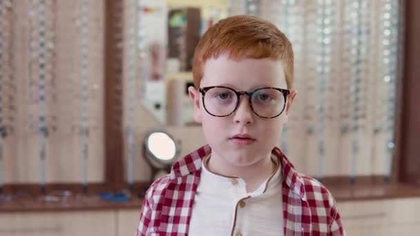 Un chico caucásico pelirrojo con una camisa a cuadros roja y blanca está de pie con gafas transparentes para la corrección de la visión y sonríe mirando directamente a la cámara — Vídeo de stock