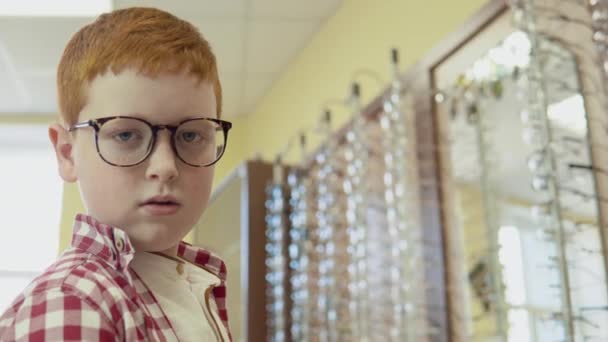 Un chico muy divertido con una camisa roja y blanca se para en la óptica con gafas para la corrección de la visión y mira con una expresión seria directamente a la cámara e imaginándose a sí mismo un chico genial — Vídeo de stock