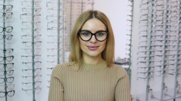 Atractiva joven mujer blanca rubia de pie delante de la cámara con gafas transparentes con un marco negro para la corrección de la visión en una tienda de gafas — Vídeos de Stock