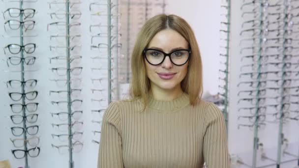 Attraktive junge blonde Frau steht in transparenter Brille mit schwarzem Rahmen zur Sehkorrektur in einem Geschäft für optische Brillen vor der Kamera und zeigt mit erhobenem Finger ein Schild — Stockvideo