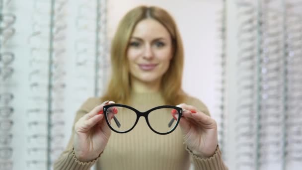 Una joven rubia caucásica sostiene anteojos delante de la cámara en una tienda de gafas y mira a la cámara. Concepto oftalmológico — Vídeos de Stock