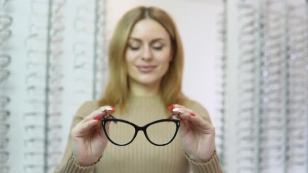 Eine blonde junge kaukasische Frau hält sich vor die Kamera, setzt dann in einem Brillengeschäft eine Sonnenbrille mit schwarzem Rand auf und blickt in die Kamera — Stockvideo