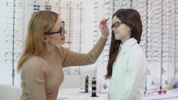 A blonde woman in a beige jacket and a dark-haired girl in a cute white cardigan and shirt pick up glasses for a childs vision correction at an eyewear store. Side view — Stock Video
