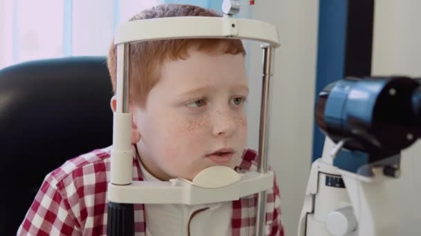 Rudowłosy biały chłopiec z aparatem ortodontycznym w kratkę w czerwono-białej koszuli w gabinecie okulistów w klinice optycznej poddaje się badaniu wzroku przy użyciu autorefraktometru. — Wideo stockowe