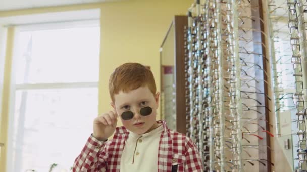 Roodharige jongen in een geruit rood-wit shirt draagt een bril tot op het puntje van zijn neus recht in de camera kijkend — Stockvideo