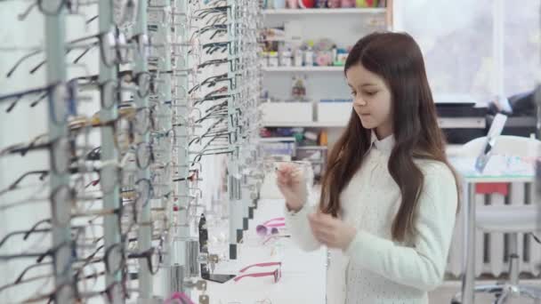 Une fille brune dans un joli cardigan blanc et chemise choisit puis porte des lunettes transparentes pour la correction de la vue dans un magasin de lunettes. Vue latérale — Video