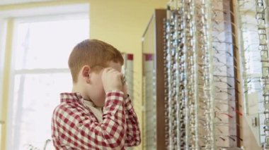 Ekose kırmızı ve beyaz tişörtlü kızıl saçlı bir çocuk mercek çerçeveli bir askılığın yanında güneş gözlüğü takar ve doğrudan kameraya bakar.