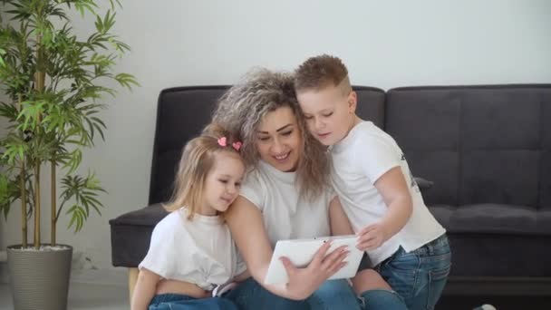 Anya, lánya és fia együtt használják a tabletet egy hangulatos nappaliban. A család ugyanabba az alapvető fehér pólóba van öltözve. Szabadidő gyerekekkel. Családi szórakoztatás — Stock videók