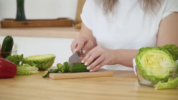 Giovane donna bionda con i capelli ricci taglia un cetriolo in anelli mentre siede a un tavolo in cucina. Vista ravvicinata di un coltello nelle mani — Video Stock