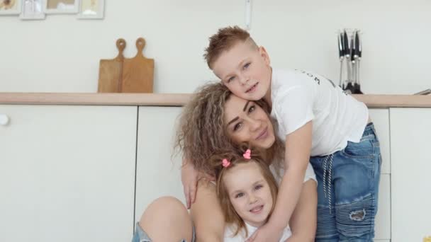 Famiglia amichevole a casa. Un ragazzo, una ragazza e una madre si siedono sul pavimento e si abbracciano in una elegante cucina moderna bianca nel design scandinavo e guardano la fotocamera. Famiglia in T-shirt e jeans bianchi — Video Stock