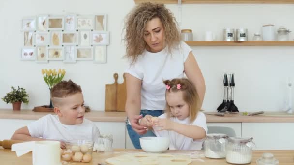 A menina quebra um ovo em uma tigela por conta própria, enquanto ela cozinha com sua mãe e irmão na cozinha em um projeto escandinavo e está envolvida em assar. Família em camisetas brancas e jeans — Vídeo de Stock