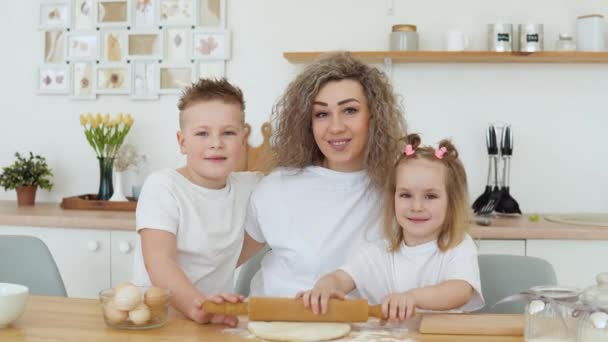 Filho, filha e mãe olham para a câmera enquanto rolam a massa juntos na cozinha. Família caucasiana europeia de loiras em camisetas brancas — Vídeo de Stock