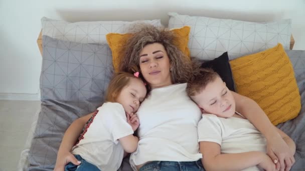 Mamá, hija e hijo yacen en la cama juntos abrazándose. Mamá y los niños están durmiendo y de repente abren los ojos y miran a la cámara. La familia está vestida con las mismas camisetas blancas básicas — Vídeos de Stock